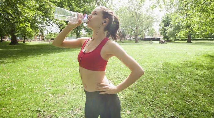Woman drinking water from a water bottle while in a park dressed in running outfit.